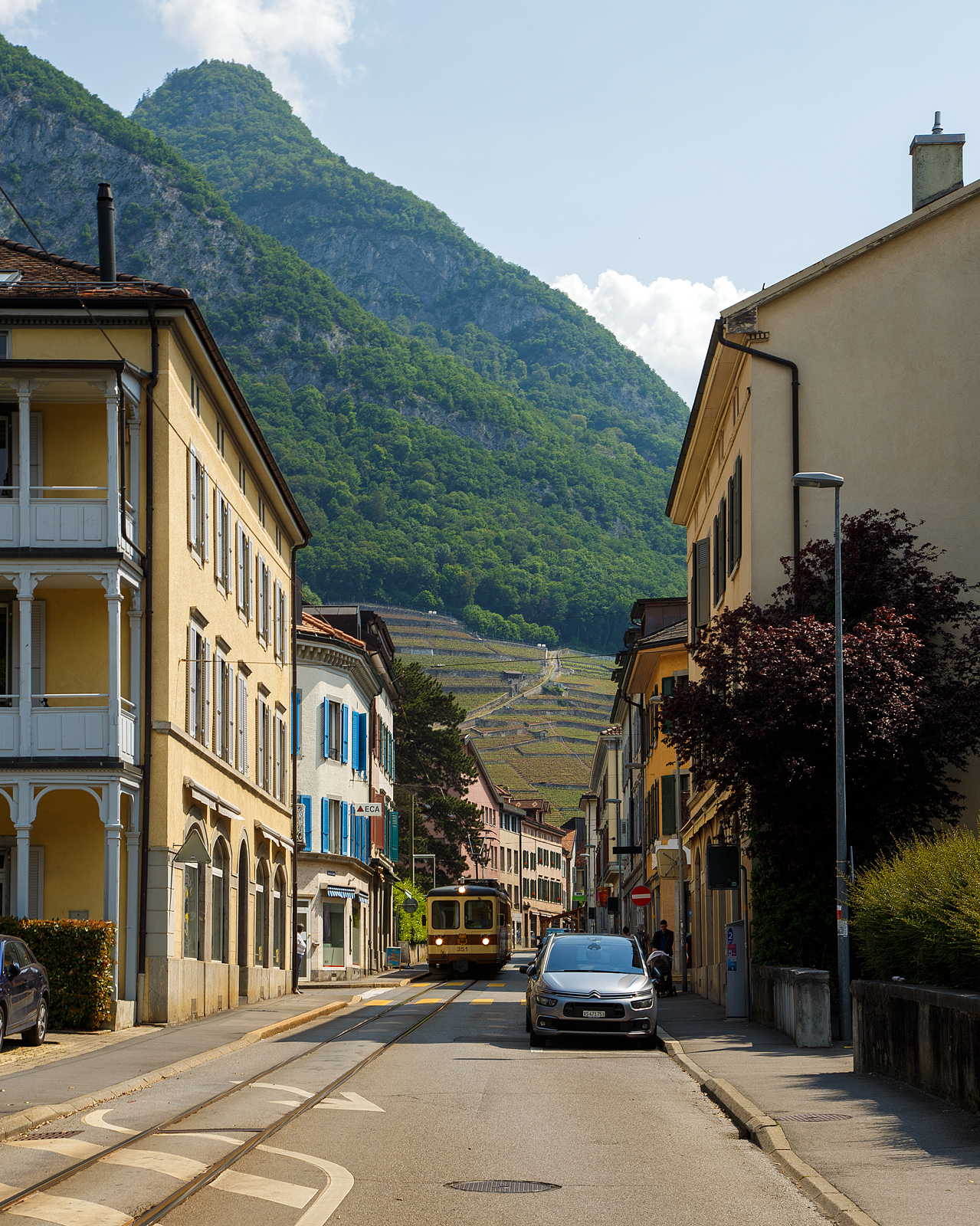 Die Straßenbahn von Aigle kommt:
Der AL Regionalzug von Leysin nach Aigle fährt am 28. Mai 2023 (hier als Straßenbahn) durch Altstadt und erreicht bald den Bahnhof Aigle. Der Zug besteht aus dem führenden Steuerwagen AL Bt 351 „Aigle“  und dem Triebwagen AL BDeh 4/4 302 „Leysin“, beide sind noch im ursprünglichen Anstrich der AL (es sind auch die einzigen). 

In Aigle (Waadt) gibt es gleich drei Schmalspurbahnen, die seit 1999 in die Transports Publics du Chablais (TPC) fusioniert worden sind, so machten wir auf unserer Rückreise hier mal einen kurzen Zwischenhalt. Die Schmalspurbahnen sind die Aigle–Ollon–Monthey–Champéry-Bahn (AOMC), die Aigle–Sépey–Diablerets-Bahn (ASD), seit dem 22. Dezember 1913 und die Aigle–Leysin-Bahn (AL).

Die Chemin de fer Aigle–Leysin (AL), deutsch Aigle-Leysin-Bahn, war eine Eisenbahngesellschaft im Schweizer Kanton Waadt. Ihre von 1900 bis 1916 eröffnete 6,2 Kilometer lange Strecke führt von Aigle im Rhonetal hinauf nach Leysin-Grand Hôtel. Die gemischte Zahnradbahn in der Spurweite 1.000 mm (Meterspur) mit dem System Abt wird seit der Betriebsaufnahme elektrisch betrieben. Im Jahr 1999 fusionierte die AL zu den Transports Publics du Chablais (TPC).

Die Bahnstrecke Aigle–Leysin hat ihren Ausgangspunkt vor dem SBB-Bahnhof und führt als Straßenbahn auf der Rue de la Gare (Bahnhofstraße), der Avenue des Ormonts und über die Grande Eau zum Kopfbahnhof Aigle-Dépôt. Dort ändern die Züge die Fahrtrichtung, damit das Triebfahrzeug für die anschließende Bergfahrt am Ende des Zuges eingereiht ist. 

Im Keilbahnhof Aigle-Dépôt beginnt der 5,3 Kilometer lange und bis zu 230 Promille steile Zahnstangenabschnitt. Zunächst führt er durch die Rebberge und bietet den Fahrgästen einen Ausblick auf das Rhonetal. Bei der Haltestelle Pont-de-Drapel wechselt die Vegetation und das Trassee befindet sich nun im Wald. Der Zug erreicht eine Waldlichtung mit dem Bahnhof Rennaz (Leysin), wo er sich in der Regel mit dem Gegenzug kreuzt. Die Strecke führt weiter durch Wald und den 154 Meter langen Tunnel Rennaz. Ab rund 1.200 Meter über Meer fährt die Zahnradbahn durch Weiden und erreicht nach kurzer Zeit den Bahnhof Leysin-Village (Leysin-Dorf). Hier beginnt der Doppelspurabschnitt mit der 128 Meter langen Brücke Leysin nach Leysin-Feydey. Nach dem 287 Meter langen Kehrtunnel Leysin erreicht die Strecke, immer noch mit Zahnstange versehen, den Endpunkt Leysin-Grand Hôtel.